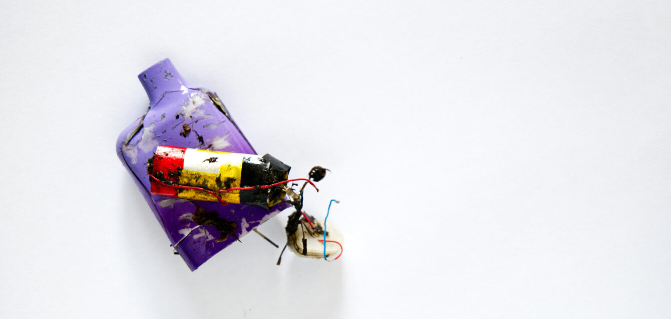 The remains of a discarded dirty muddy neon coloured electronic cigarette vape, and its internal components and battery. Shot above a white paper background.